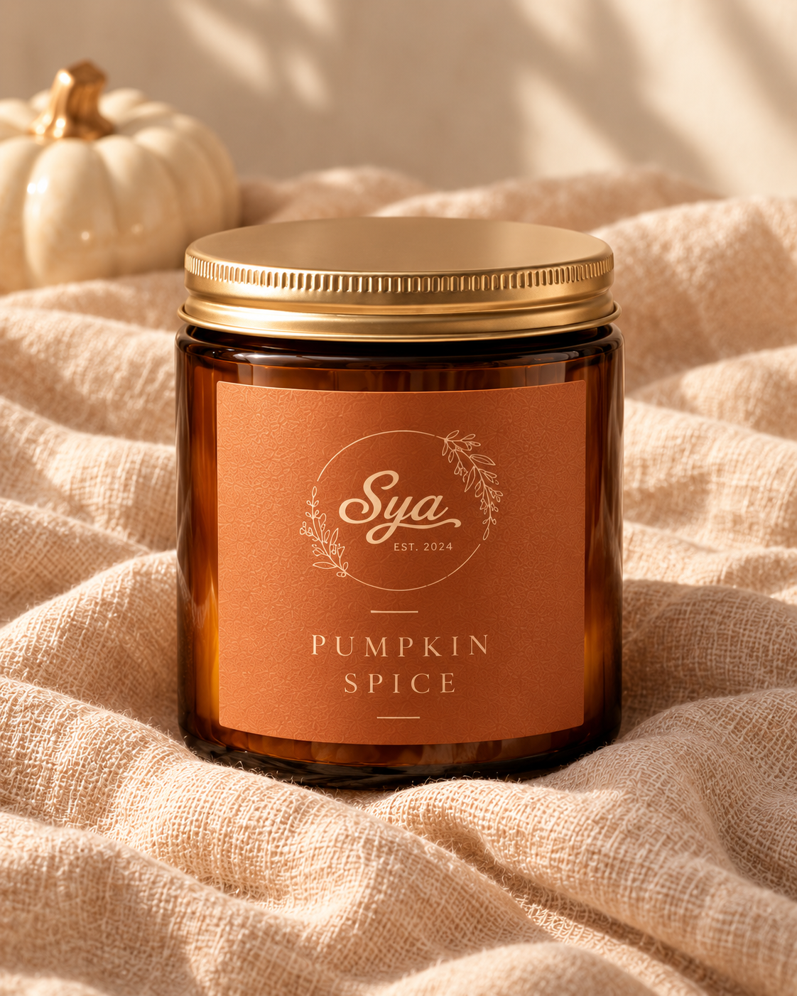 Vela Sya en frasco de vidrio ámbar con tapa dorada y etiqueta naranja Pumpkin Spice, sobre tela beige junto a una calabaza decorativa.
