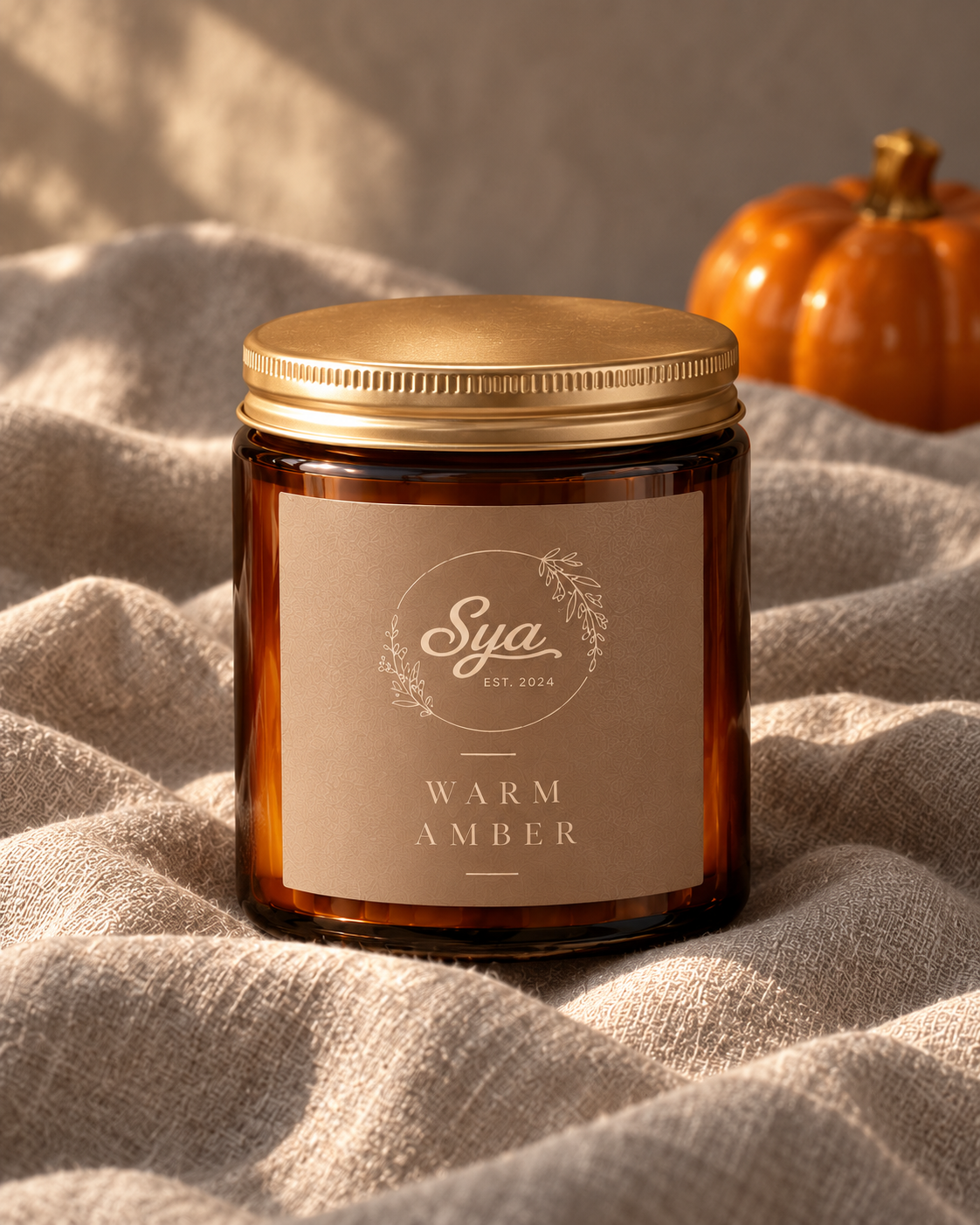 Vela Sya en frasco ámbar con tapa dorada y etiqueta beige Warm Amber, sobre tela con una calabaza naranja decorativa al fondo.
