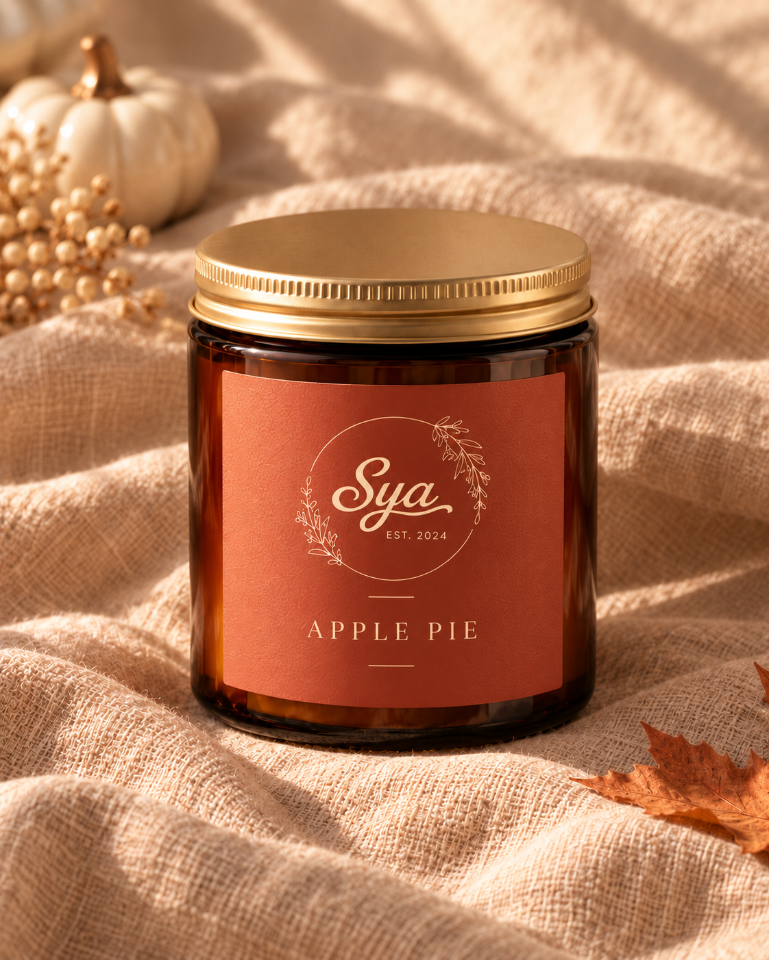 Vela Sya en frasco ámbar con tapa dorada y etiqueta naranja Apple Pie, sobre tela beige con flores secas y hojas otoñales.