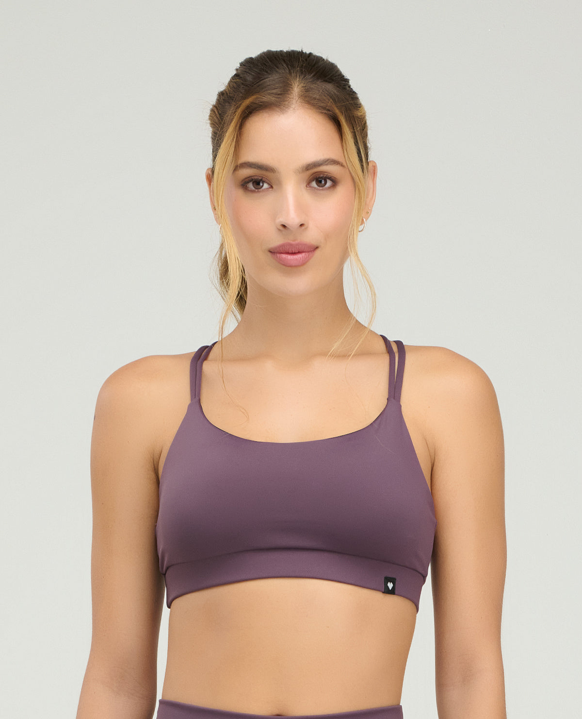 Mujer usando top deportivo morado de silueta ajustada con copas suaves y cargaderas dobles sobre fondo gris.