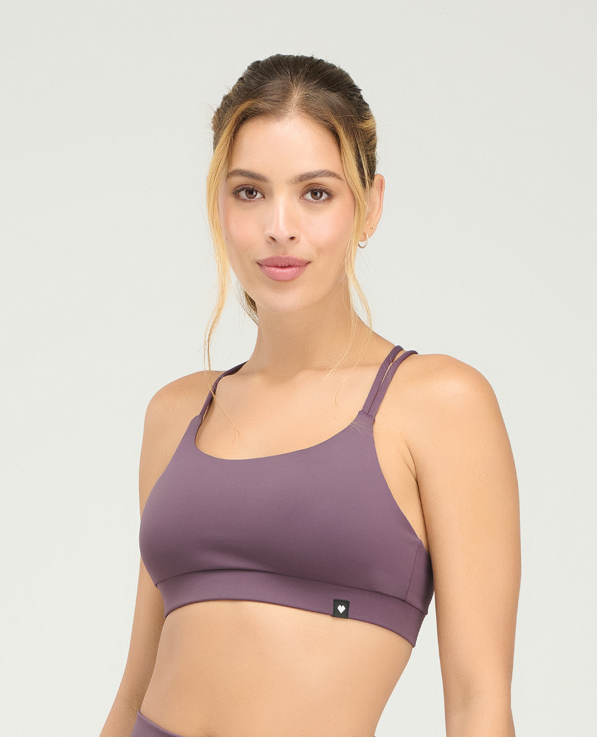 Top deportivo color morado para mujer con copas fijas suaves y cargaderas dobles sobre fondo gris claro.