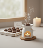 White ceramic oil burner with smoke rising, surrounded by chocolates and a lit candle on a wooden coaster.